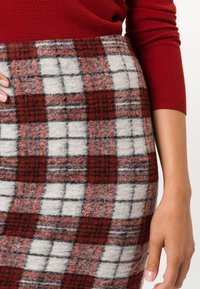Jupe en laine avec un motif tartan rouge, blanc et noir. Design ajusté avec un tissu texturé. Main reposant sur le côté gauche.