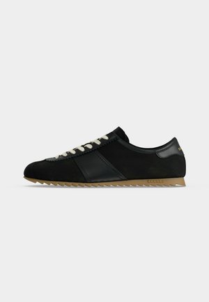 Zwarte lage sneaker met witte veters, gestructureerde bruine zool en leren details tegen een effen grijze achtergrond.