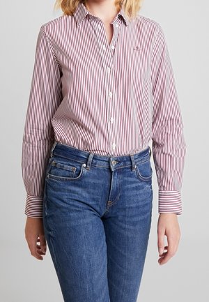 Personne portant une chemise à manches longues rouge et blanche à rayures verticales, rentrée dans un jean en denim bleu, sur un fond uni.