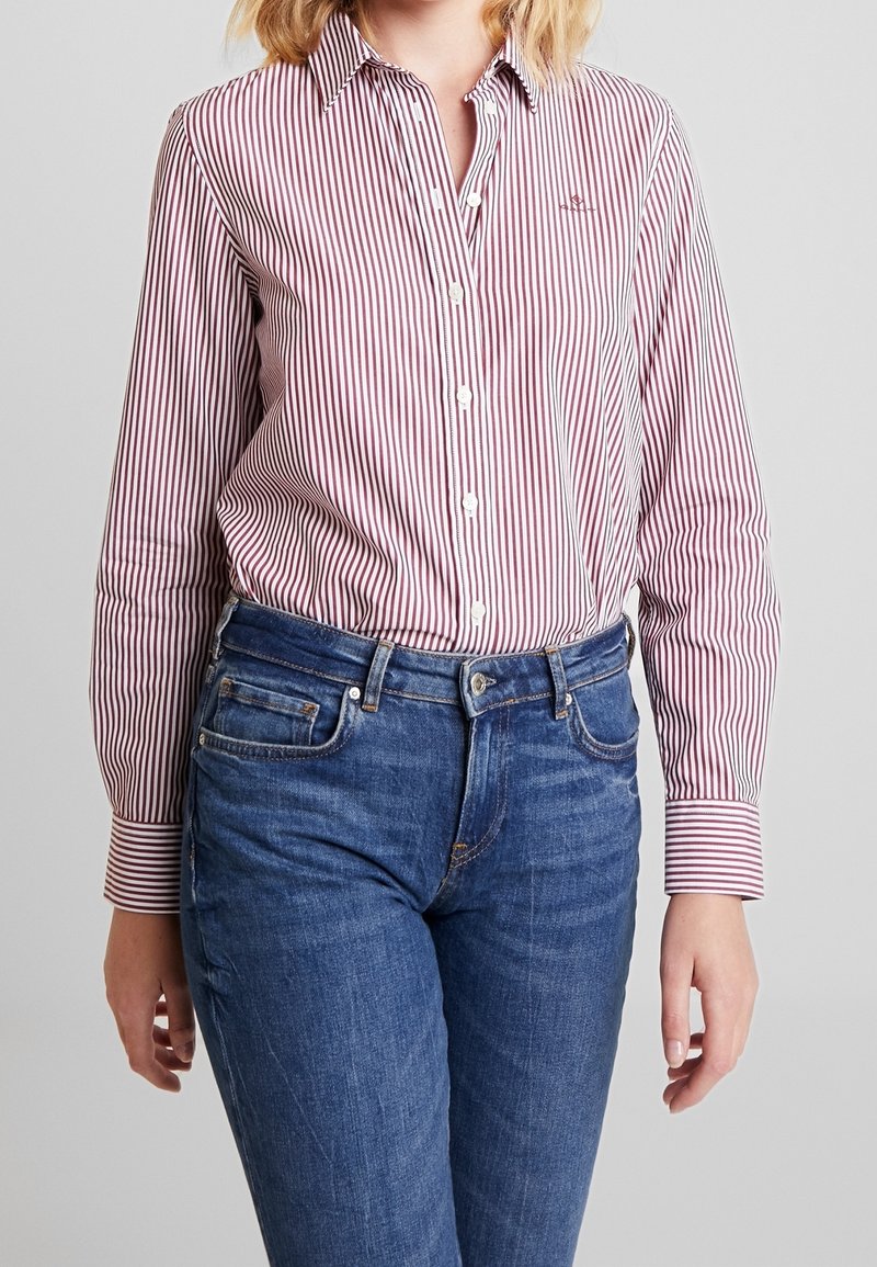Personne portant une chemise à manches longues rouge et blanche à rayures verticales, rentrée dans un jean en denim bleu, sur un fond uni.