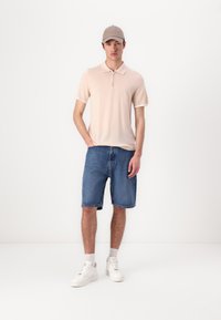 Světle růžové polo tričko se zipovým límcem, spárované s modrými denimovými šortkami a bílými teniskami. Design obsahuje kontrastní bílé akcenty.