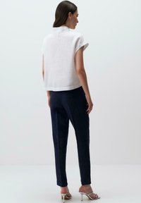 Weißes Leinen-T-Shirt mit kurzen Ärmeln, kombiniert mit marineblauen strukturierten Hosen. Das Model trägt silberne High Heel Sandalen. Der Hintergrund ist einfarbig.