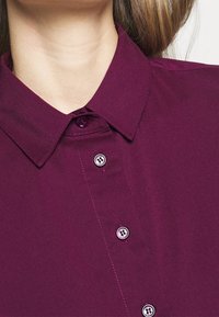 Camicia collare viola con placca a bottoni e bottoni tondi, tessuto liscio e dettagli di cucitura sottili sul colletto.