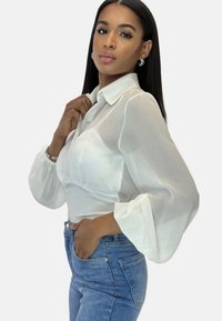 Blouse blanche transparente avec un col haut, des manches longues bouffantes et un corsage ajusté. Portée avec un jean en denim bleu clair, orné de poches.