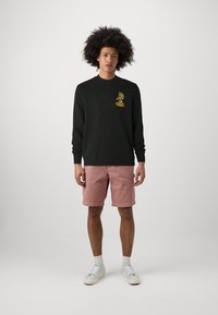 Zwarte sweatshirt met gele grafische print op de borst, gecombineerd met roze shorts. Model draagt witte sneakers en sokken, staand tegen een lichte achtergrond.