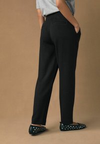 Person wearing black tailored pants and black studded flats, standing with hands in pockets against a plain brown background.