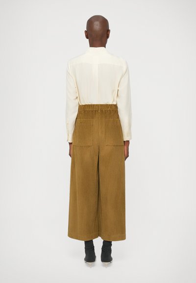 Camisa color crema con mangas con puños, combinada con pantalones acampanados de pana marrón que tienen una cinturilla elástica y dos bolsillos traseros.