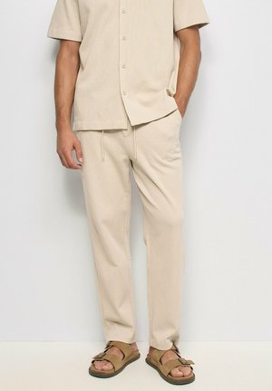 Hombre con camisa de manga corta beige, pantalones con cordón a juego y sandalias marrones de punta abierta, de pie frente a un fondo claro y sencillo.