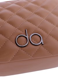 Bolso de mano acolchado en color beige, hecho de faux cuero suave, que presenta un acento de logo metálico oscuro y detalles de costura visibles.
