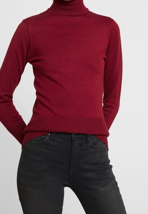 Person trägt einen eng anliegenden, langärmeligen roten Rollkragenpullover und schwarze Jeans mit hoher Taille, steht mit den Händen hinter dem Rücken.