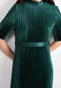 Plissé jurk in donkergroene satijn, met brede korte mouwen, een hoge hals en een bijpassende ceintuur in de taille. Verticale plooien over het geheel.