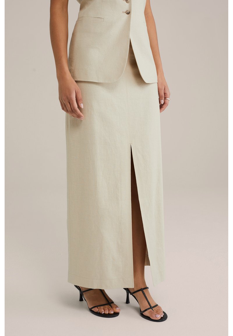 Beige linnen maxi rok met een split aan de voorkant, rechte vorm en schone naden. Gecombineerd met zwarte sandalen met bandjes. Formeel ontwerp geschikt voor evenementen.