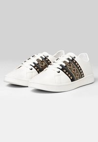 Zapatillas blancas con cordones, con franjas laterales de patrón geométrico en negro y oro con remaches, y suelas redondas blancas.