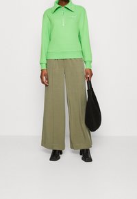 Zelená mikina na zip s límcem, spárovaná s širokými olivovými kalhotami a černými kotníkovými botami. Nese měkkou černou tašku.