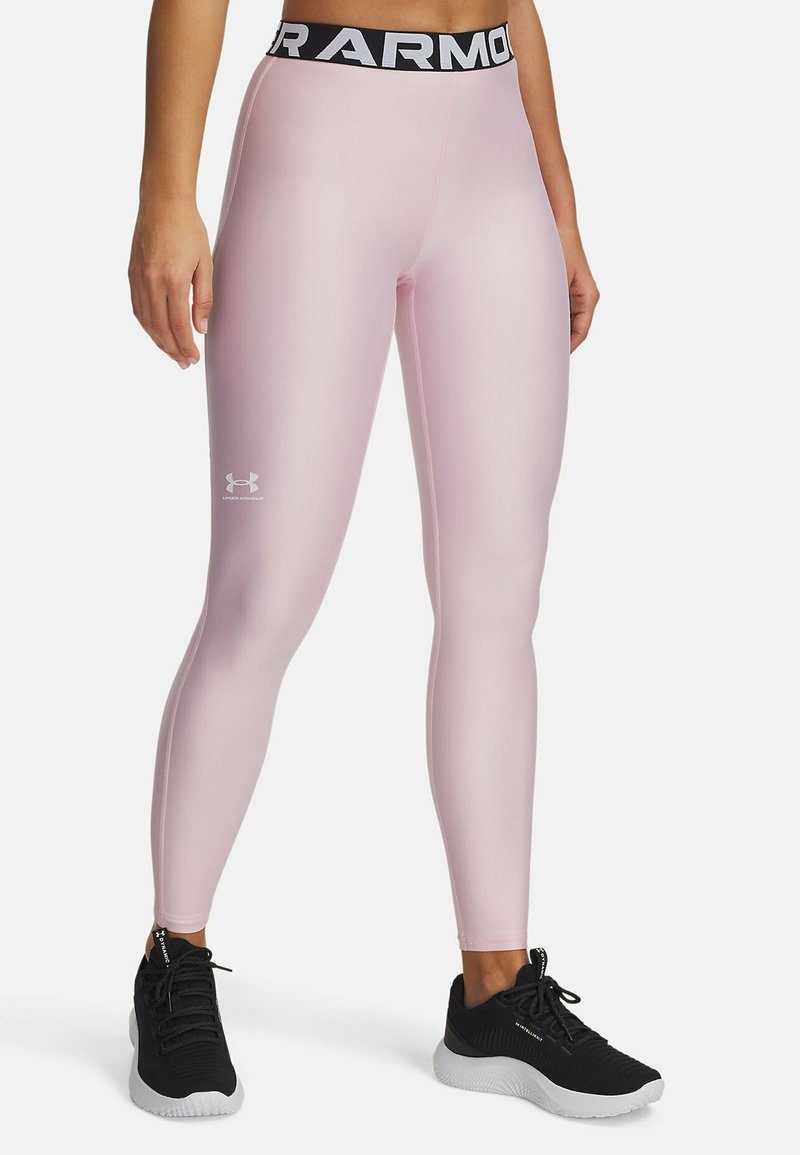 Svetlo roza športne legice z črnim pasom, na katerem je napis "Under Armour". So tesno prilegajoče in imajo gladko, sijočo teksturo.