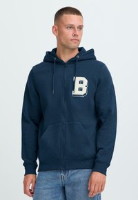 Mladý muž s krátkymi vlasmi, ktorý má na sebe námornícku modrú mikinu na zips s bielym písmenom "B" na hrudi a svetlomodré džínsy.