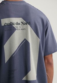 Asmuo, vilkintis mėlyną marškinėlį su dideliais baltais geometriniais raštais ir prancūzišku užrašu "étoile du Nord" atspausdintu ant nugaros.