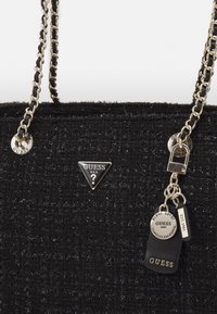 Czarna torebka z tweedu z metalowym łańcuszkiem, srebrnym okuciem i wieloma metkami. Wyraźne logo na trójkątnej plakietce. Teksturowane wykończenie.
