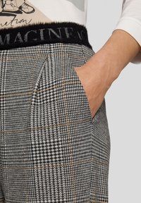 Hand in der Tasche einer hoch taillierten grauen Karohose mit schwarzem, flauschigem Bund, der teilweise den bestickten Schriftzug "IMAGINE" zeigt.