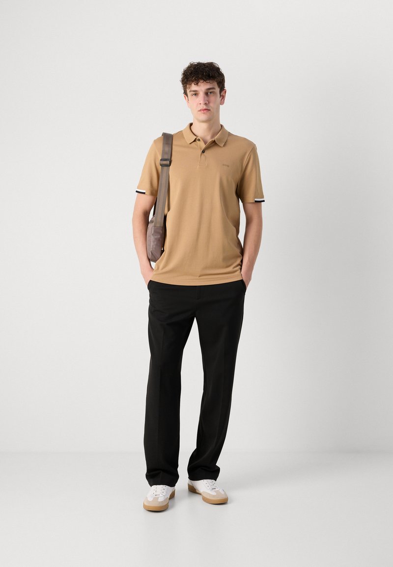 Polo shirt tan avec des poignets rayés noir et blanc, accompagné de pantalons noirs et de baskets blanches. Le mannequin tient un sac bandoulière marron.