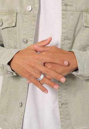 Anello di metallo con una superficie texturizzata, indossato su una mano. La persona indossa una giacca di jeans verde chiaro sopra una camicia bianca.