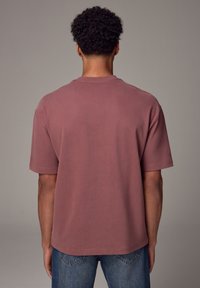 T-shirt in cotone bordeaux con vestibilità rilassata, maniche corte e scollatura a girocollo, abbinato a jeans in denim blu. Nessun motivo o dettaglio evidente.