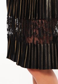 Svart plisserad kjol med guldaccenter, med en spetsöverdrag vid fållen. Texturerad tyg som skapar en lager-på-lager effekt.