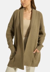 Frau trägt eine lockere, grob gestrickte olivgrüne Strickjacke mit strukturierten Mustern, vorderen Taschen und einem breiten, gerippten Schal-Kragen.