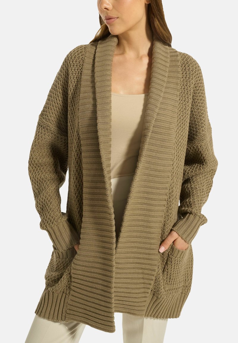 Frau trägt eine lockere, grob gestrickte olivgrüne Strickjacke mit strukturierten Mustern, vorderen Taschen und einem breiten, gerippten Schal-Kragen.