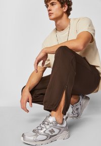 Adidași argintii din material mesh și sintetic, asociați cu pantaloni de jogging maro închis cu fermoare la gleznă și un tricou bej deschis.