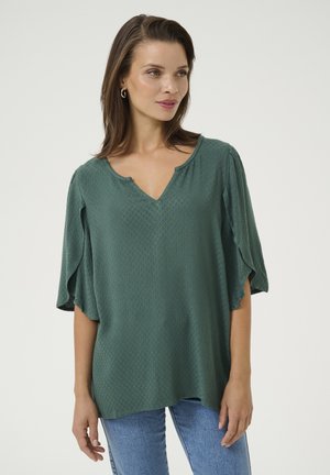 Haut vert texturé avec un décolleté en V et des manches évasées, associé à un jean bleu clair. Motif de losanges notable sur le tissu.
