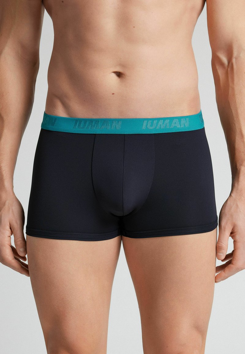 Boxer briefs noirs en tissu lisse, avec une ceinture élastique bleu sarcelle portant la marque « IUMAN », longueur mi-cuisse, design minimaliste.