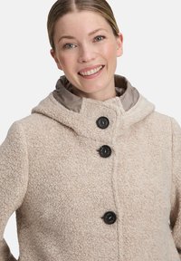 Beige comfortabele fleecjas met een capuchon, voorzien van zwarte knopen, een gestructureerde stof en een gladde binnenvoering in een contrasterende kleur.