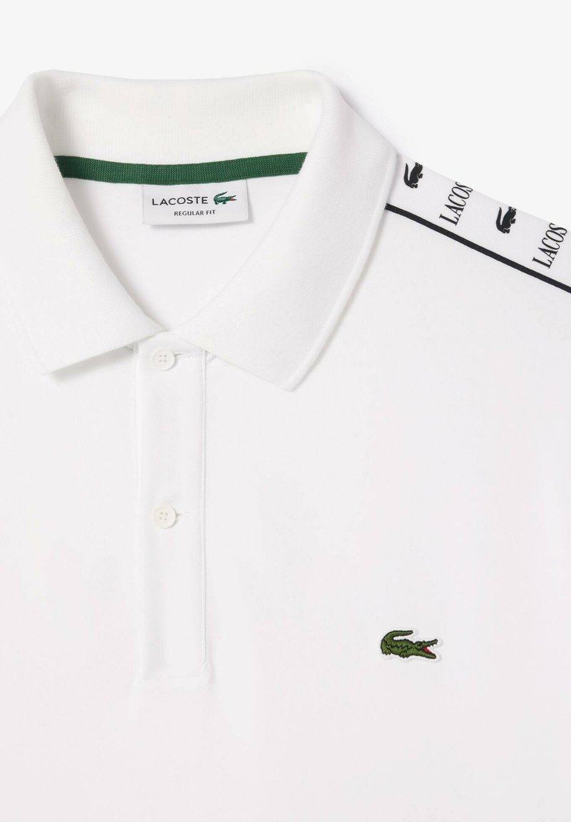 Polo shirt Lacoste blanc avec logo crocodile vert sur la poitrine et bandelette de marque le long de l'épaule, col à regular fit avec deux boutons.