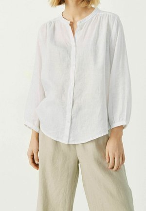 Kvinde iført en løs hvid knap-op bluse med rynket halsudskæring og beige bukser med vide ben mod en ensartet lys baggrund.