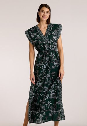 Lächelnde Frau, die ein dunkelgrünes Maxikleid mit Blumenmuster und gemustertem Besatz trägt, V-Ausschnitt, Kappenärmel und seitlichem Schlitz, hält den Saum mit einer Hand.