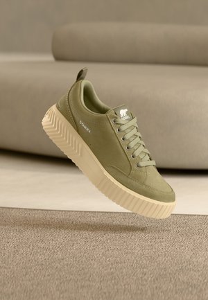 Sneaker verde oliva chiaro con lacci e suola spessa beige, con marchio "Sorel" sul lato e sulla linguetta, mostrata su uno sfondo di tessuto beige.