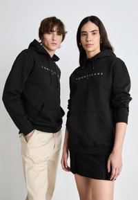 Sudaderas negras con el logo "TOMMY JEANS" en dorado, con un bolsillo canguro y cordones, llevadas por dos personas sobre un fondo neutro.