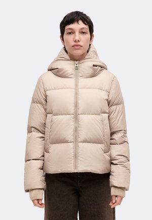 Personne aux cheveux courts et foncés portant une doudoune beige à col montant et fermeture éclair à l'avant, associée à un pantalon foncé.