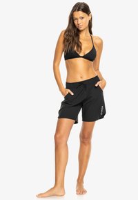 Haut de bikini noir avec des bretelles fines, associé à un short de bain noir ample avec une taille élastique et un détail logo sur la jambe.