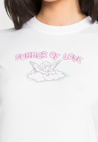 Bílý bavlněný tričko s grafickým designem, na kterém je nápis "SUMMER OF LOVE" v růžové barvě, zobrazující cheruba na oblaku s obrysem křídel.