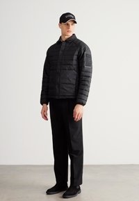 Fekete pufi dzseki modern kialakítással, texturált felülettel és patentgombokkal, fekete, lapos elejű nadrággal és sapkával párosítva.