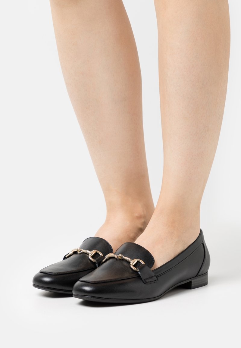 Pieds chaussés de mocassins en cuir noir avec un détail en métal argenté, posés sur une surface blanche.