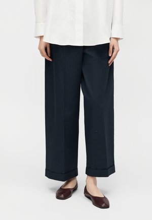 Oseba, oblečena v temno mornarsko modre široke hlače z zavihanimi robovi, belo srajco z dolgimi rokavi in temno rjave nizke čevlje na belem ozadju.