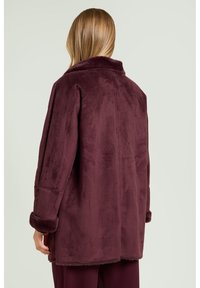 Bordeauxfarvet imiteret pelsjakke med krave, lange ærmer og blød tekstur. Bemærkelsesværdige detaljer inkluderer bløde ærmekanter og et afslappet pasformdesign.