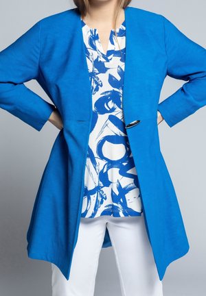 Femme portant une veste bleu vif ouverte sur un chemisier blanc avec des motifs abstraits bleus et un pantalon blanc, mains sur les hanches.