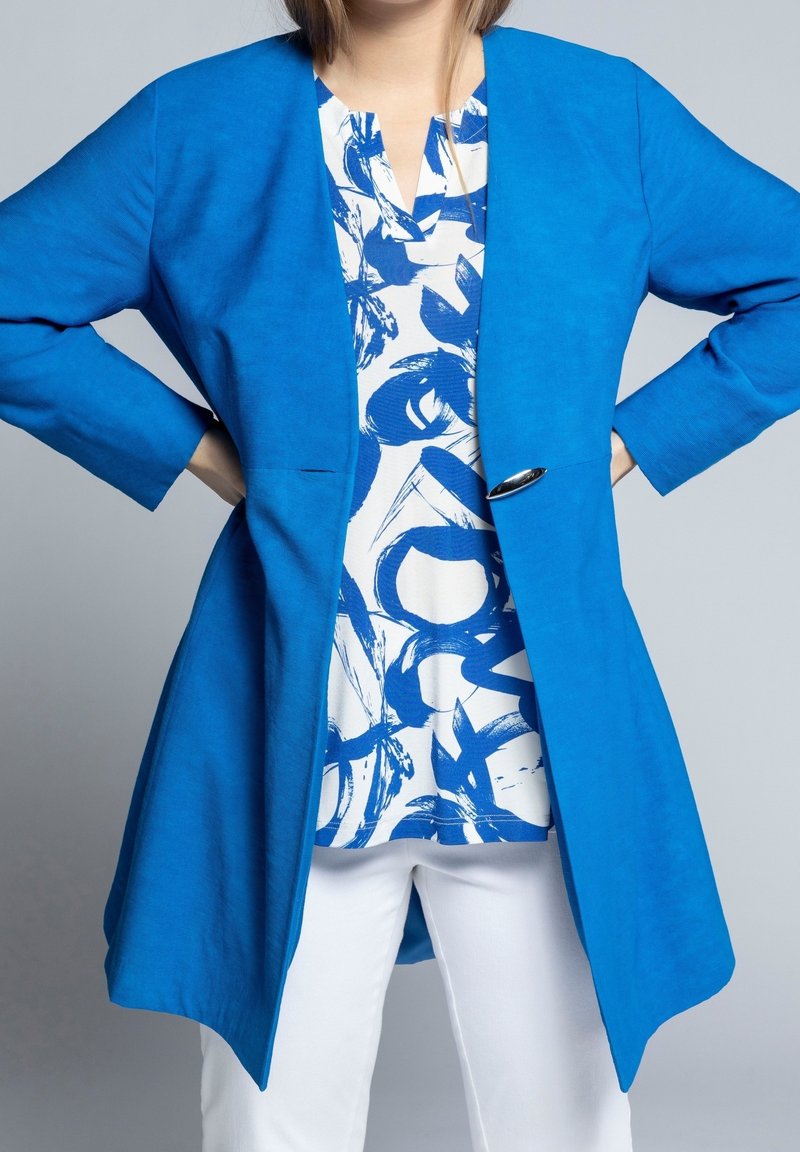 Femme portant une veste bleu vif ouverte sur un chemisier blanc avec des motifs abstraits bleus et un pantalon blanc, mains sur les hanches.