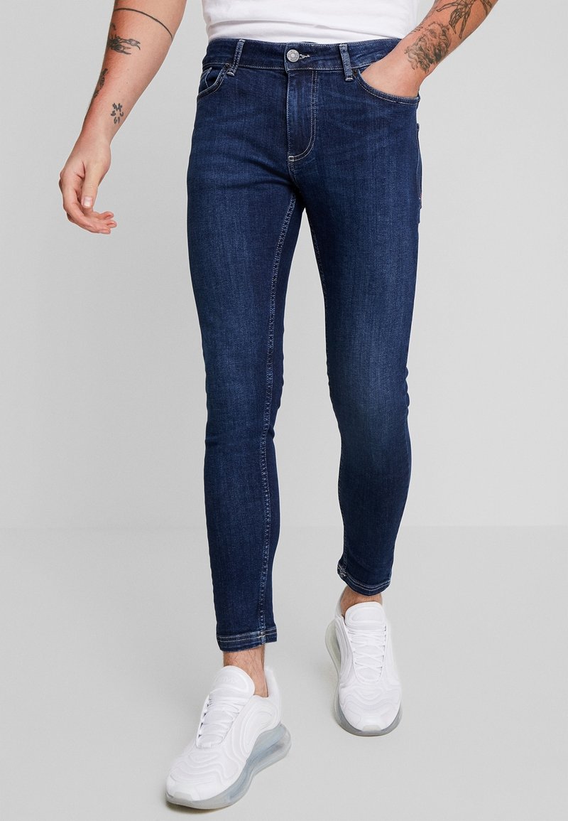 Jeans ajustados de un azul oscuro, hechos de denim, que cuentan con cinco bolsillos, cierre de botón y costuras visibles como acento. Combinados con zapatillas blancas.