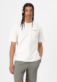 T-shirt en coton crème à manches courtes, avec des graphiques "McLaren" et "MONACO" en texte sombre et orange. Un short olivier texturé.