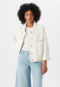 Fehér denim dzseki laza szabással, nagy gallérral és ezüst gombokkal, világoskék bő szárú farmernadrággal és fehér inggel párosítva.